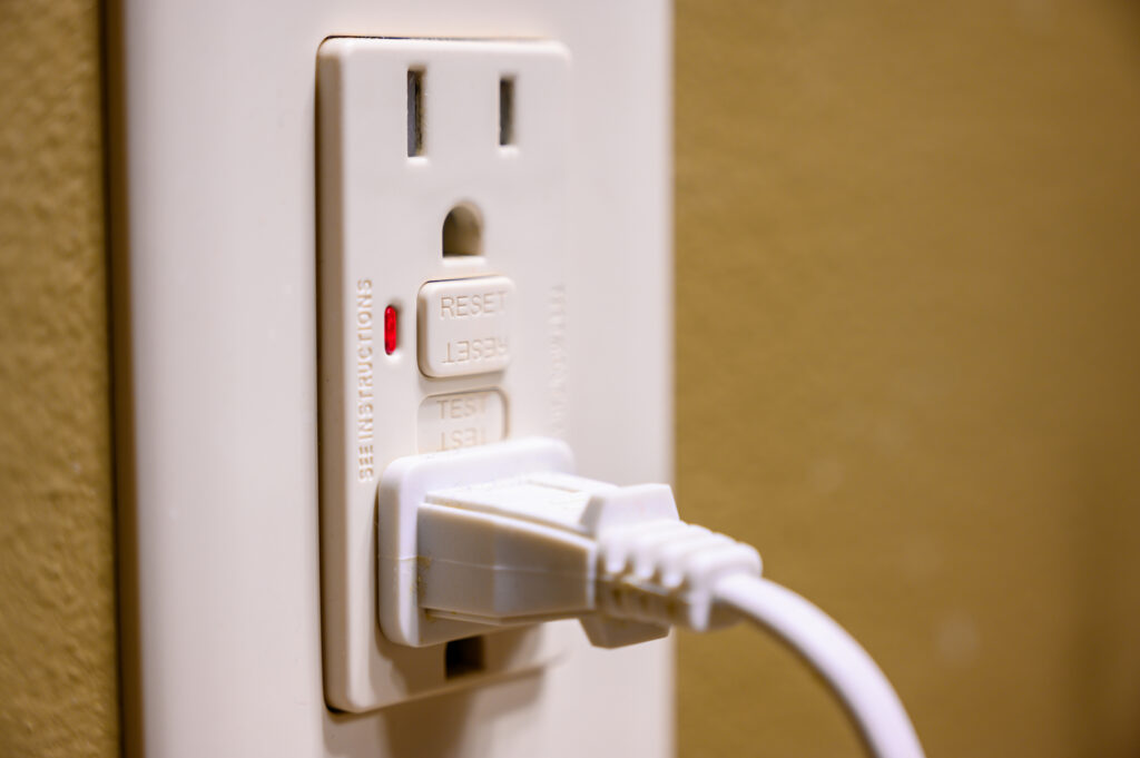 Residential electrical outlet with a cord plugged into the bottom outlet, a reset button between the two outlets, on a mustard-yellow wall.