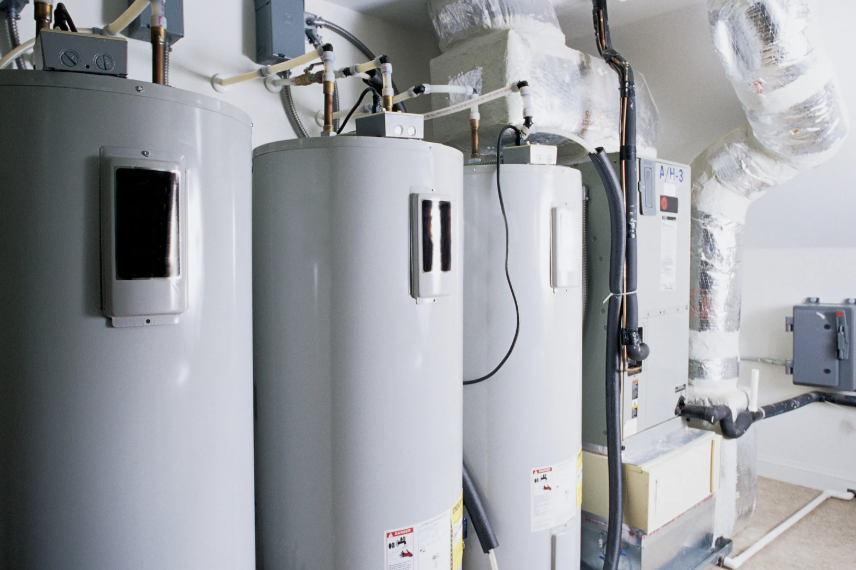 Three large gray water heaters stand side by side in a utility room, surrounded by insulated pipes, vents, and electrical panels.