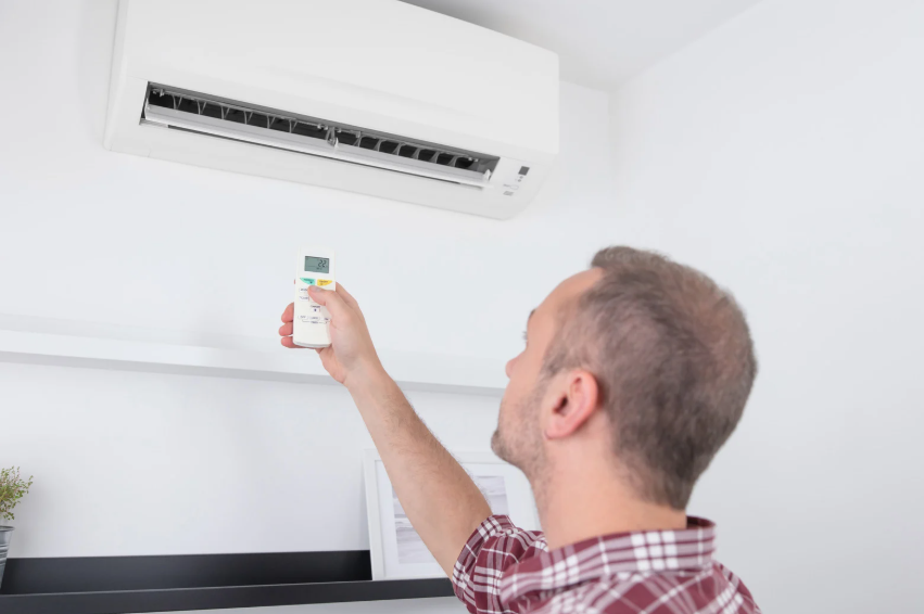 A man in a plaid shirt uses a remote control to operate a wall-mounted air conditioner in a bright room.