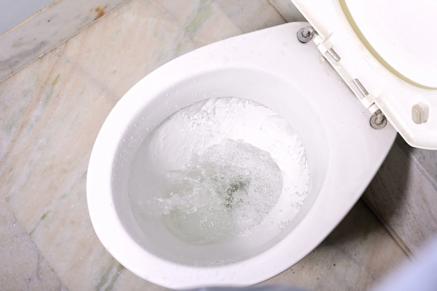 A white toilet with the lid up is flushing, showing swirling water inside the bowl. The toilet is on a light-colored tiled floor.