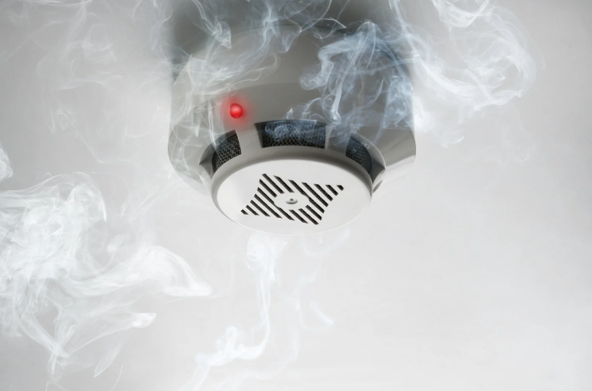 Ceiling-mounted smoke detector with a red warning light activated, surrounded by visible smoke.