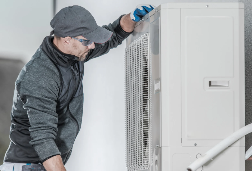 A technician wearing a cap, sunglasses, and gloves inspects or repairs an outdoor air conditioning unit mounted on a wall.