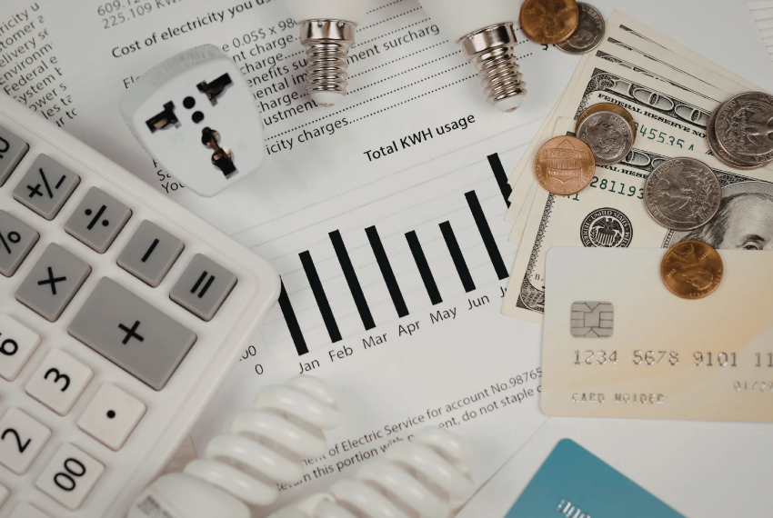 Calculator, utility bills, light bulbs, coins, cash, and a credit card are arranged on a table, suggesting household budgeting and energy expenses.