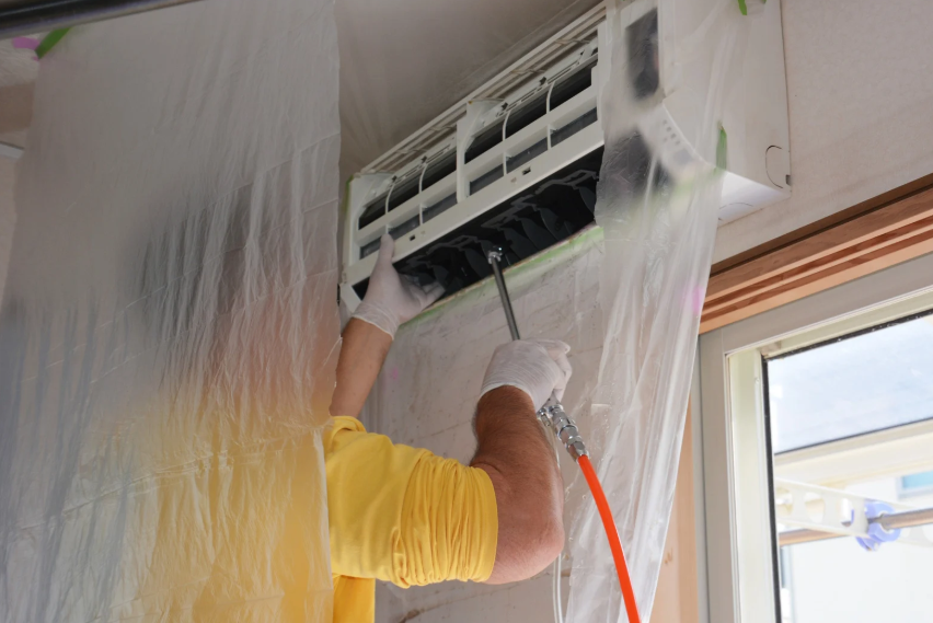 A person in a yellow shirt and gloves cleans an air conditioner unit using a spray tool, with plastic sheeting covering the surrounding area.