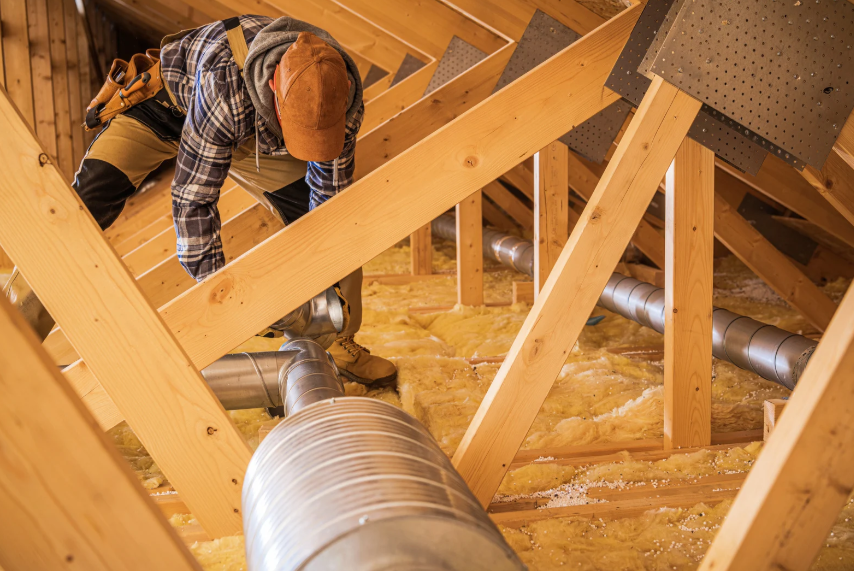 A worker in a flannel shirt and hard hat installs metal ductwork in an unfinished attic with exposed beams and insulation.