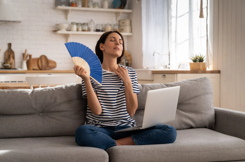 A woman sits cross-legged on a couch with a laptop on her lap, fanning herself with a blue hand fan in a bright kitchen or living room.