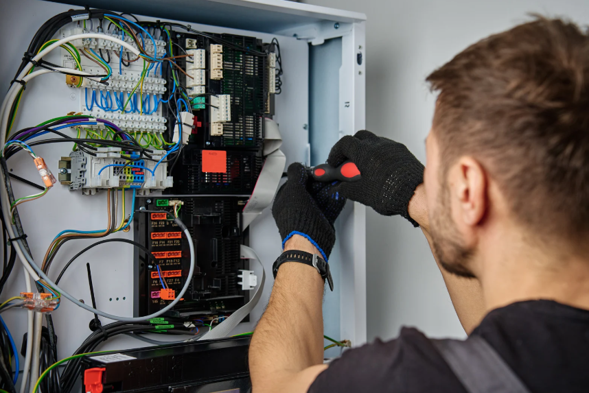A person wearing black gloves uses a screwdriver to work on an open electrical control panel with various wires and components.