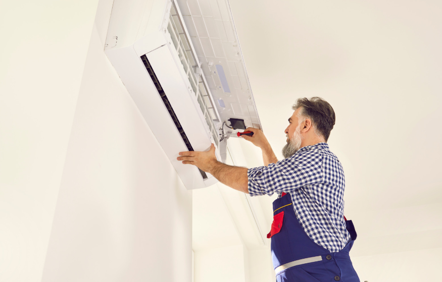 A man in work overalls uses a screwdriver to repair or maintain a wall-mounted air conditioning unit.