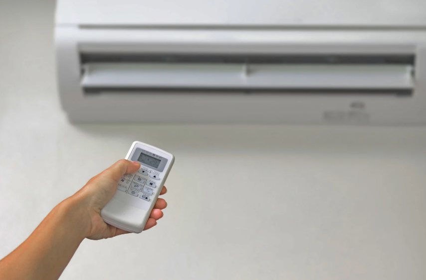 A hand holds a remote control pointed at a wall-mounted air conditioner unit, preparing to adjust its settings.