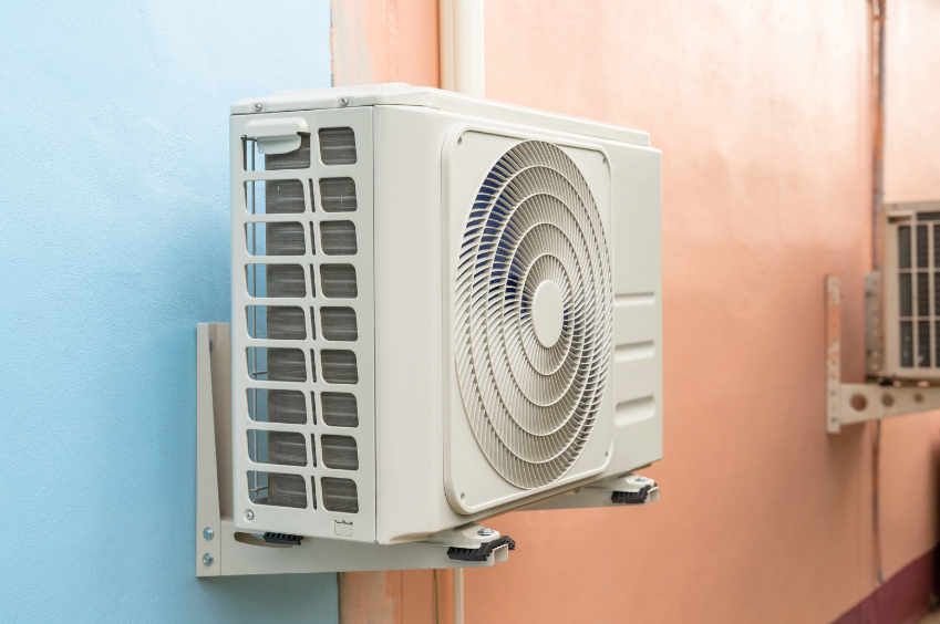 A white outdoor air conditioning unit is mounted on a blue wall next to a peach-colored wall. Another unit is visible in the background.