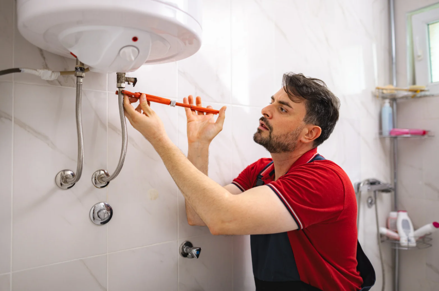 A person in a red shirt uses a wrench to adjust pipes connected to a wall-mounted water heater in a tiled bathroom.