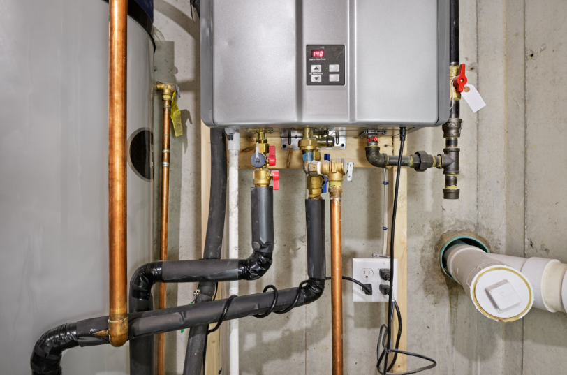 A tankless water heater with connected copper pipes and valves mounted on a basement wall, next to other plumbing and electrical components.
