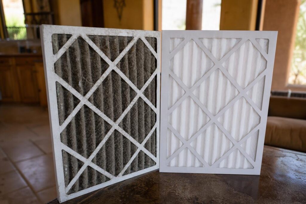 A dirty air filter stands next to a clean air filter on a table in a room with tan walls and windows in the background.