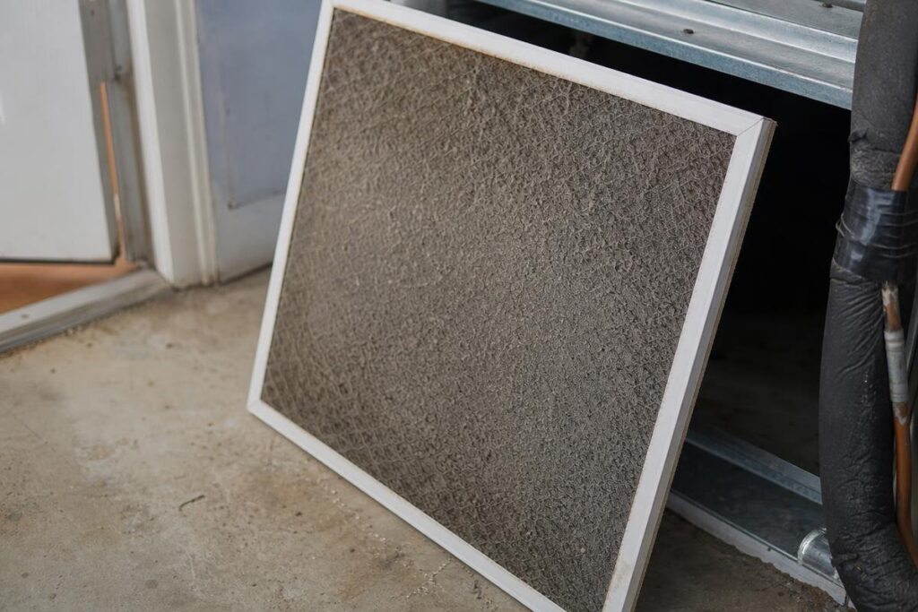 A dirty air filter leaning against an HVAC unit on a concrete floor, with visible dust and debris on the filter surface.